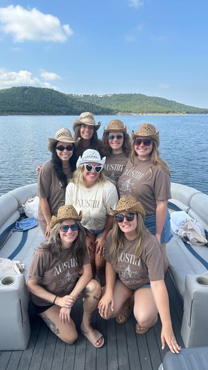 Eight women in straw cowboy hats and matching "Austin" shirts smiling together on a pontoon boat with a calm lake, tree-covered hills and blue sky in the background.