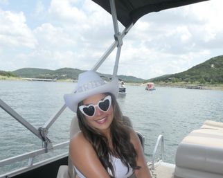 Smiling woman wearing a BRIDE cowboy hat and rhinestone heart-shaped sunglasses relaxing on a pontoon boat on a sunny lake with rolling green hills in the background
