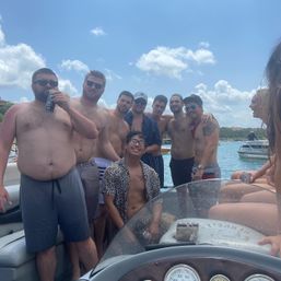 Group of men in swimwear enjoying a boat party on a sunny summer day, posing with drinks on a motorboat with blue sky, scattered clouds and other boats in the background