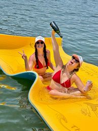 Two friends in red bikinis relaxing on a yellow floating mat on a lake, one pouring champagne from a bottle while the other flashes a peace sign, both wearing sunglasses and caps on a sunny summer day.