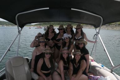 Group of friends in swimsuits and straw cowboy hats smiling and posing on a covered pontoon boat on a scenic lake with wooded shoreline — summertime boating fun