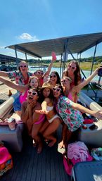 Cheerful group of women on a pontoon boat at a sunny lake dock, wearing swimsuits, floral shirts and heart-shaped sunglasses for a fun summer boat party.
