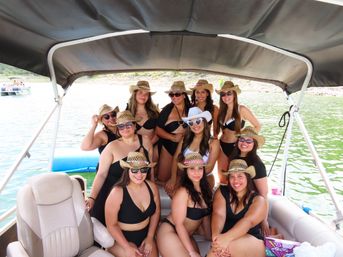 Group of friends in black swimsuits and straw cowboy hats smiling together on a covered pontoon boat during a sunny lake day with a rocky shoreline