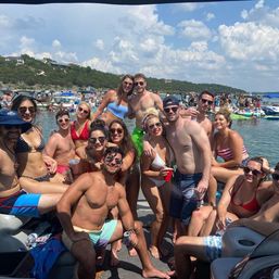 Smiling group of young adults in swimsuits posing on a crowded pontoon boat at a sunny lake party, holding drinks with other boats, inflatables and a tree‑covered shoreline under a blue sky.