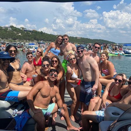 Smiling group of young adults in swimsuits posing on a crowded pontoon boat at a sunny lake party, holding drinks with other boats, inflatables and a tree‑covered shoreline under a blue sky.