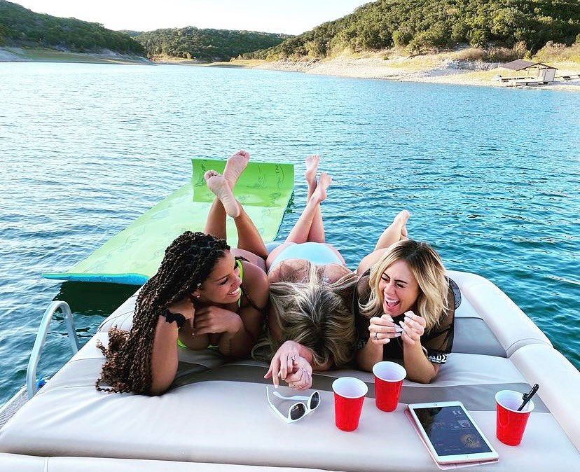 Three friends lounging on the bow of a boat over a blue lake, laughing on a green float mat with red cups, sunglasses and a tablet nearby and a wooded shoreline in the background