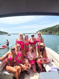 Eight friends in pink bikinis posing on a pontoon boat at a sunny lake — bachelorette-style boat party with paddleboarders and tree-lined shoreline in the background.