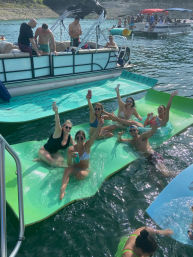 Friends enjoying a summer lake party on bright green floating mats beside pontoon boats, raising drinks and smiling.