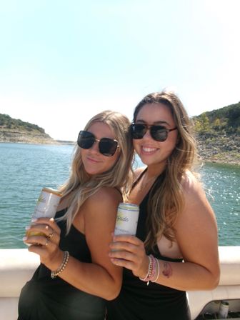Two friends on a boat enjoying a sunny lake day, wearing black swimsuits and sunglasses and holding canned drinks with rocky shoreline behind.