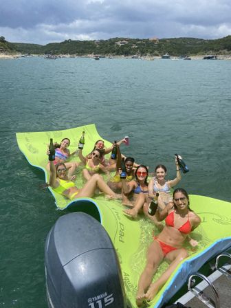 Group of friends in colorful swimsuits lounging on a lime-green floating pad beside a boat, toasting with bottles and cans on a sunny summer lake with wooded shoreline and boats in the distance.