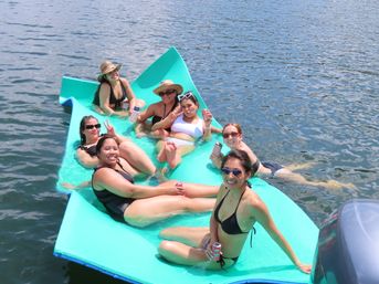 Seven women in swimsuits relaxing on a bright turquoise floating raft on a lake, wearing sunglasses and sun hats and holding canned drinks on a sunny summer day.