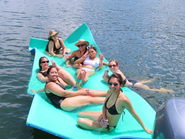 Seven women in swimsuits relaxing on a bright turquoise floating raft on a lake, wearing sunglasses and sun hats and holding canned drinks on a sunny summer day.