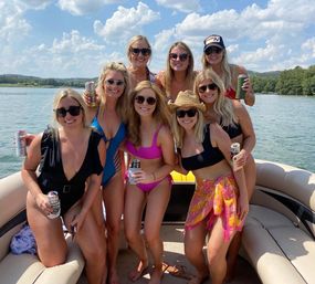 Eight friends in swimsuits smiling and holding canned drinks on a pontoon boat on a sunny lake, blue sky with fluffy clouds and tree-lined shore in the background