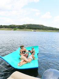 Four friends in swimsuits relaxing on a bright turquoise floating mat on a calm lake, smiling and posing with sunglasses and a straw hat, with a tree-lined shoreline and hills with homes in the background under a partly cloudy sky.