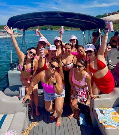 Group of smiling women in colorful swimsuits and pink hats cheering with drinks on a pontoon boat at a sunny lake