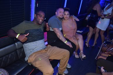 Three friends posing in a blue-lit party bus lounge on a night out — a man giving a thumbs-up, a tattooed man with his arm around a woman in a pink dress and high heels, with other partygoers and LED lighting in the background.