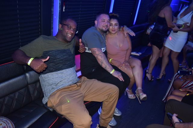 Three friends posing in a blue-lit party bus lounge on a night out — a man giving a thumbs-up, a tattooed man with his arm around a woman in a pink dress and high heels, with other partygoers and LED lighting in the background.