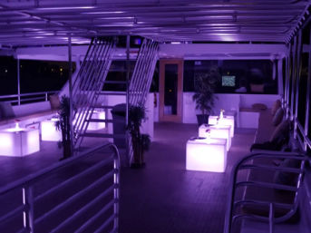 Purple-lit yacht upper deck lounge at night with glowing cube tables, cushioned bench seating, metal stairway, potted plants and railing