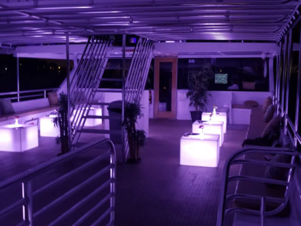 Purple-lit yacht upper deck lounge at night with glowing cube tables, cushioned bench seating, metal stairway, potted plants and railing