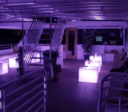 Purple-lit yacht upper deck lounge at night with glowing cube tables, cushioned bench seating, metal stairway, potted plants and railing