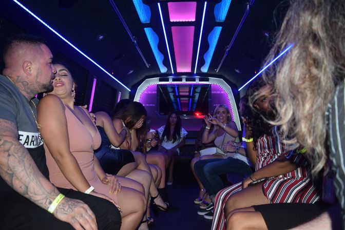 Friends partying inside a neon-lit party limo with pink and blue LED ceiling lights, guests seated on leather benches laughing, posing and taking photos.