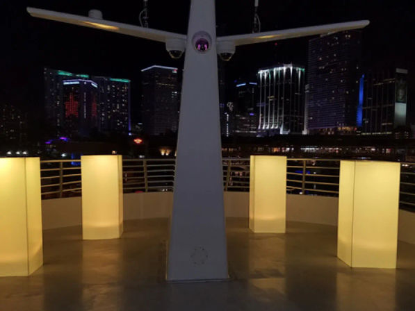 Night rooftop terrace with four glowing yellow rectangular light pillars around a central white modern sculpture, illuminated city skyline of high-rise buildings in the background.