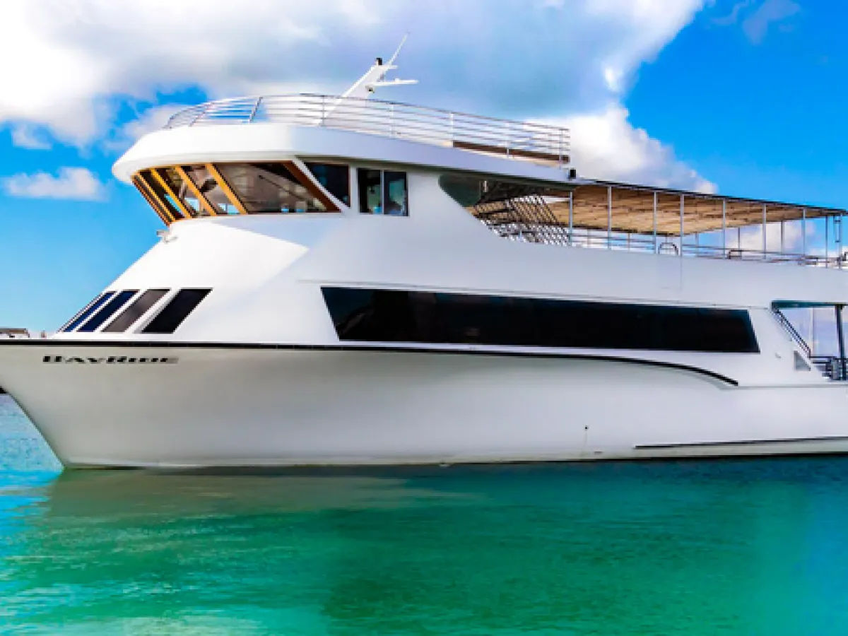 Sleek white luxury yacht floating on clear turquoise water under a bright blue sky with puffy clouds, showing panoramic windows and an upper sun deck.