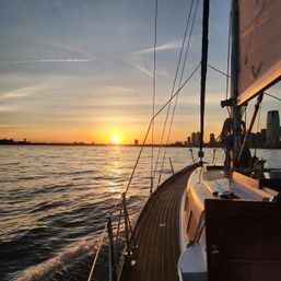 Golden sunset reflected on rippling water, view from a wooden-deck sailboat with rigging in the foreground and a distant city skyline on the horizon