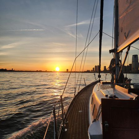 Golden sunset reflected on rippling water, view from a wooden-deck sailboat with rigging in the foreground and a distant city skyline on the horizon