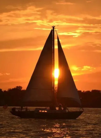 Sailboat silhouette with tall triangular sails gliding across shimmering water at golden sunset, the sun glowing through the sails against a tree-lined horizon.
