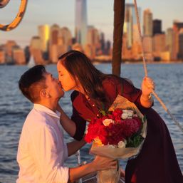 Romantic couple kissing on a sailboat at sunset, woman holding a vibrant red and white bouquet with a golden city skyline across the water.