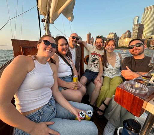 Six friends smiling and holding canned drinks on a sailboat at sunset, with an urban waterfront skyline in the background.