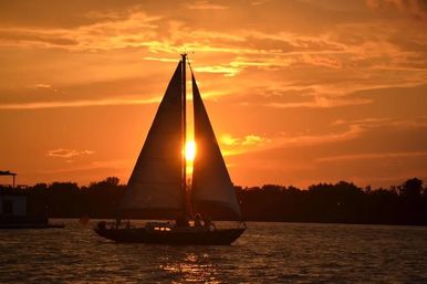 Sailboat silhouette at sunset with the sun glowing between its sails, golden-orange sky and reflective water beneath a tree-lined horizon.