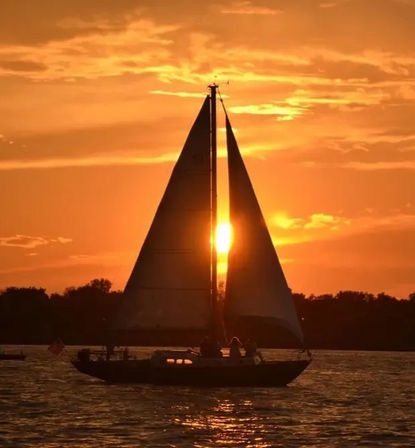 Sailboat silhouette on a tranquil lake at golden sunset, bright orange sky with the sun perfectly framed between the sails
