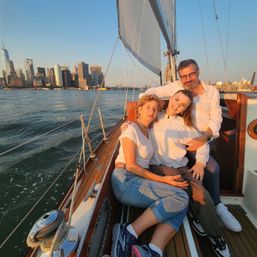 Three smiling people cuddled on a sailboat at golden hour with the Manhattan skyline and NYC waterfront in the background