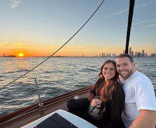 Smiling couple seated on a sailboat at sunset, holding a drink with the Manhattan skyline and Statue of Liberty visible across sparkling water