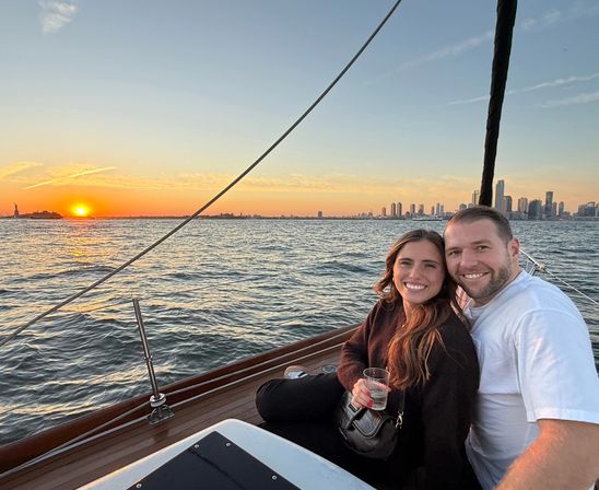 Smiling couple seated on a sailboat at sunset, holding a drink with the Manhattan skyline and Statue of Liberty visible across sparkling water