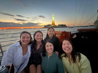 Five friends smiling on a sailboat at sunset in New York Harbor, with the illuminated Statue of Liberty on Liberty Island and a colorful skyline on the horizon