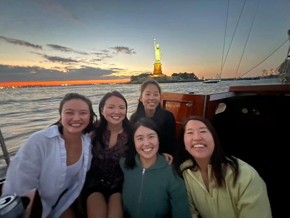 Five friends smiling on a sailboat at sunset in New York Harbor, with the illuminated Statue of Liberty on Liberty Island and a colorful skyline on the horizon