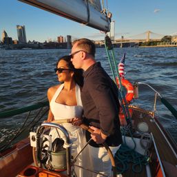 Couple steering a sailboat at golden hour on New York's East River with the Manhattan skyline and Brooklyn Bridge in the background, American flag on deck.