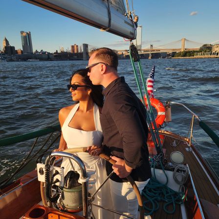 Couple steering a sailboat at golden hour on New York's East River with the Manhattan skyline and Brooklyn Bridge in the background, American flag on deck.