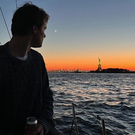 Silhouetted person on a sailboat gazing at the Statue of Liberty across New York Harbor at sunset with a crescent moon and rippling water in the foreground.