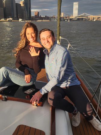 Smiling couple on a sailboat at golden hour, woman showing an engagement ring with the Manhattan skyline and Brooklyn Bridge in the background