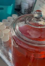 Close-up of a glass beverage dispenser filled with chilled red punch, condensation on the jar and glass lid knob, with stacked white sample cups on a silver tray nearby — event drink station.