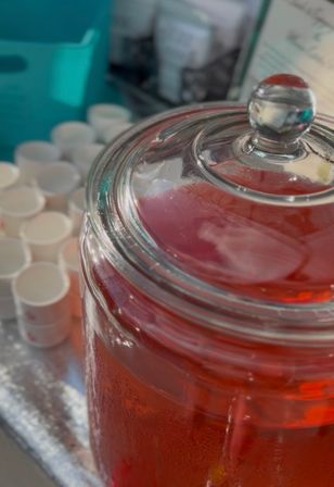 Close-up of a glass beverage dispenser filled with chilled red punch, condensation on the jar and glass lid knob, with stacked white sample cups on a silver tray nearby — event drink station.