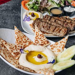 Close-up of Mediterranean mezze: creamy hummus drizzled with olive oil and topped with an olive and edible flower, served with toasted pita triangles, cucumber slices, and a plate of grilled kofta, mixed greens and dipping sauces.