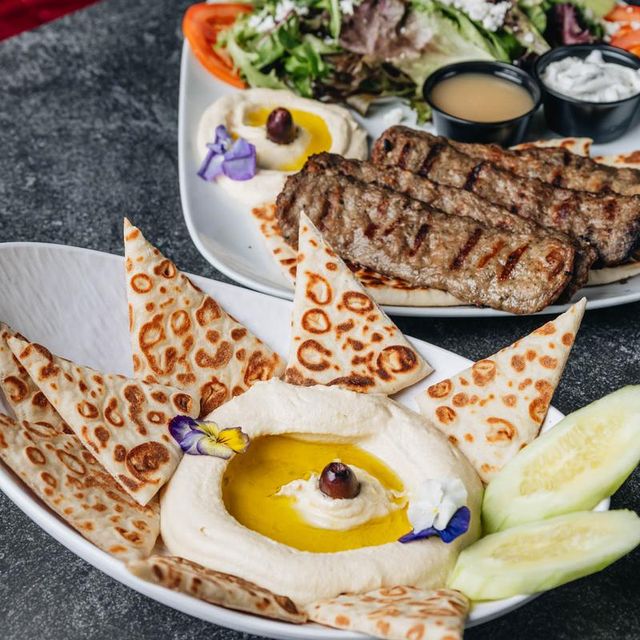 Close-up of Mediterranean mezze: creamy hummus drizzled with olive oil and topped with an olive and edible flower, served with toasted pita triangles, cucumber slices, and a plate of grilled kofta, mixed greens and dipping sauces.