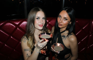 Two people toasting with rose-gold coupe cocktails in a red tufted booth, wearing black outfits with visible tattoos during a dimly lit night out