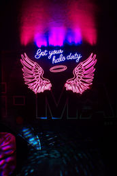 Pink and blue neon sign in a dark lounge: glowing angel wings and halo with cursive text 'Get your halo dirty', colorful reflections on wall and floor