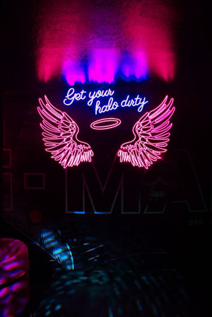 Pink and blue neon sign in a dark lounge: glowing angel wings and halo with cursive text 'Get your halo dirty', colorful reflections on wall and floor